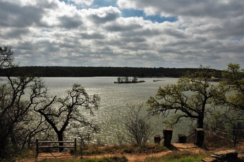 A serene lake surrounded by trees under a cloudy sky, with a small island visible in the distance.