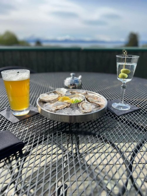 A plate of oysters with lemon, a glass of beer, and a cocktail with olives on a table with a scenic view.
