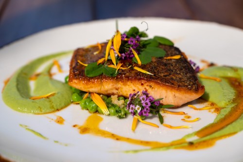 A beautifully plated salmon fillet garnished with herbs and edible flowers, served with green puree and sauce.