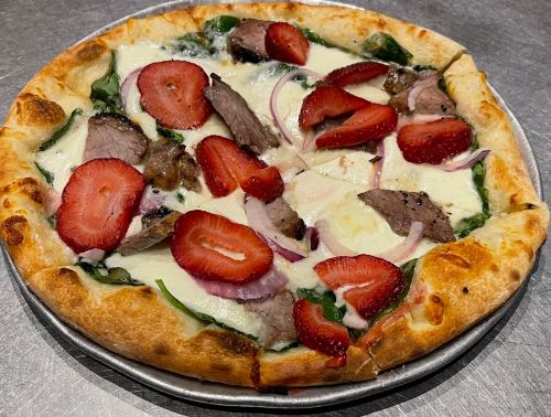 A pizza topped with spinach, sliced strawberries, grilled meat, red onions, and creamy cheese on a golden crust.
