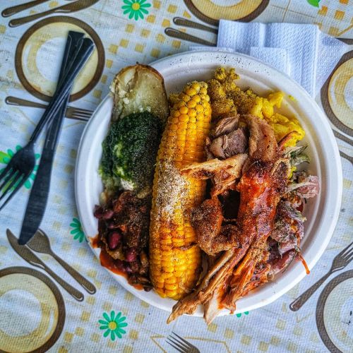 A plate of food featuring corn, broccoli, beans, a baked potato, and seasoned pulled meat on a patterned tablecloth.