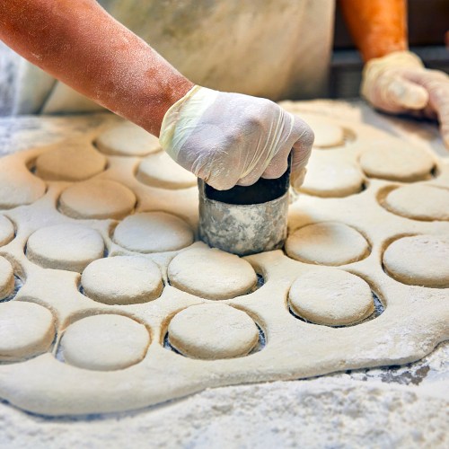 A hand in a glove presses a metal cutter into dough, creating circular shapes on a floured surface.