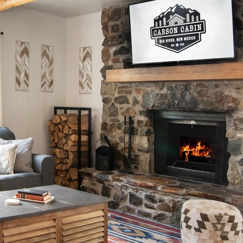 Cozy living room with a stone fireplace, TV, and rustic decor, featuring a wood stack and colorful rug.