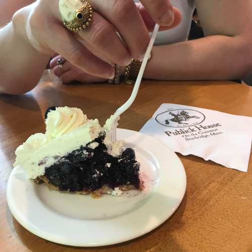 A hand holding a fork, poised to take a bite of a slice of blueberry pie topped with whipped cream on a plate.