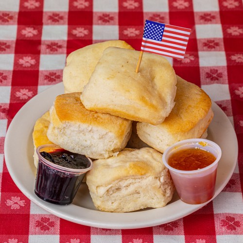 A plate of fluffy biscuits with a small American flag, accompanied by jelly and syrup on a red checkered tablecloth.