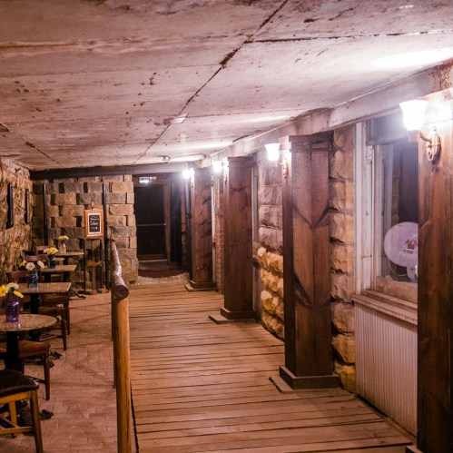 A dimly lit corridor with stone walls, wooden beams, and small tables, creating a rustic, cozy atmosphere.