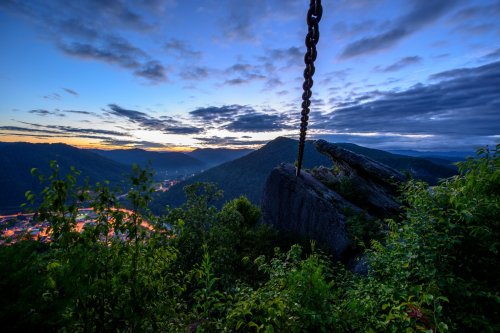A scenic view of mountains at dusk, featuring a large chain hanging from a rock, with a valley illuminated below.