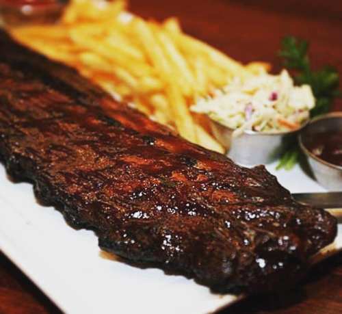 A plate featuring a large, glazed rib rack, crispy fries, coleslaw, and two small bowls of barbecue sauce.