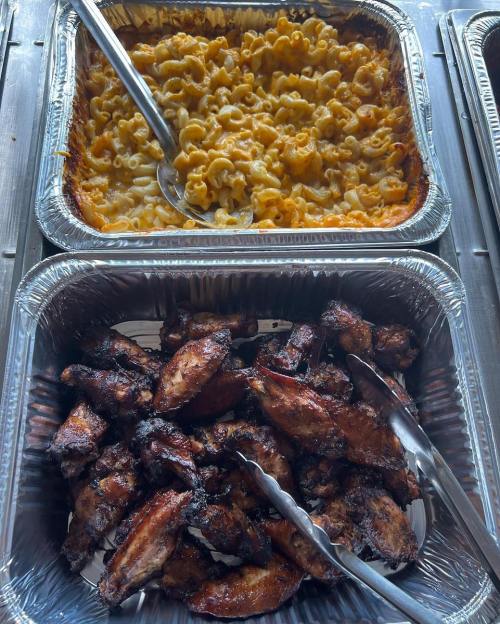 A tray of baked macaroni and cheese above a tray of grilled chicken wings, both served with tongs.