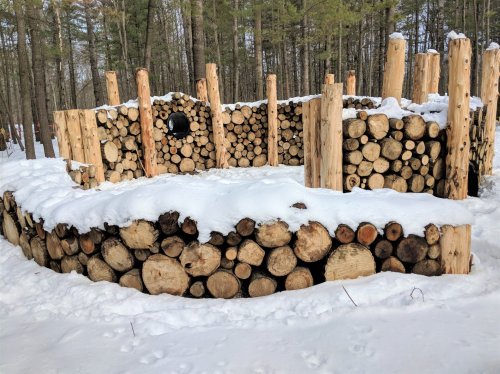 A circular structure made of stacked logs, partially covered in snow, surrounded by trees in a winter landscape.
