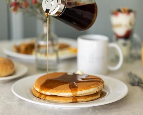 A stack of pancakes drizzled with syrup, with a coffee mug and a parfait in the background on a dining table.