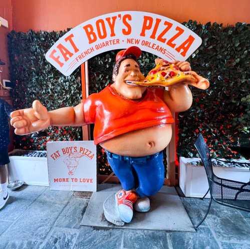 A large, cartoonish statue of a man in a red shirt eating pizza, standing in front of a "Fat Boy's Pizza" sign.