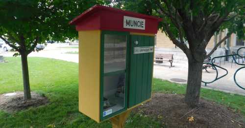 A colorful little free library with a red roof and green doors, located in a grassy area near trees.