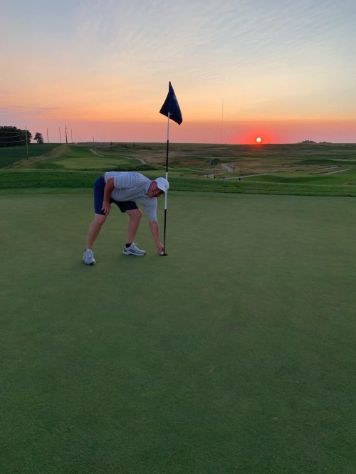 A golfer retrieves the ball from the hole at sunset, with a flag and a scenic landscape in the background.