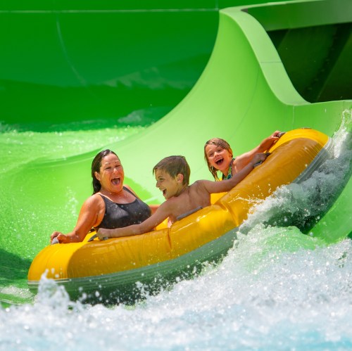 A woman and two children joyfully ride a yellow inflatable raft down a water slide, splashing in bright green water.