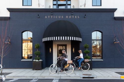 Two people ride bicycles in front of the Atticus Hotel, featuring a stylish entrance and decorative plants.