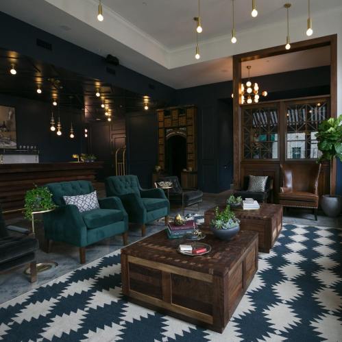 A stylish lounge with dark walls, modern furniture, and warm lighting, featuring green chairs and a patterned rug.