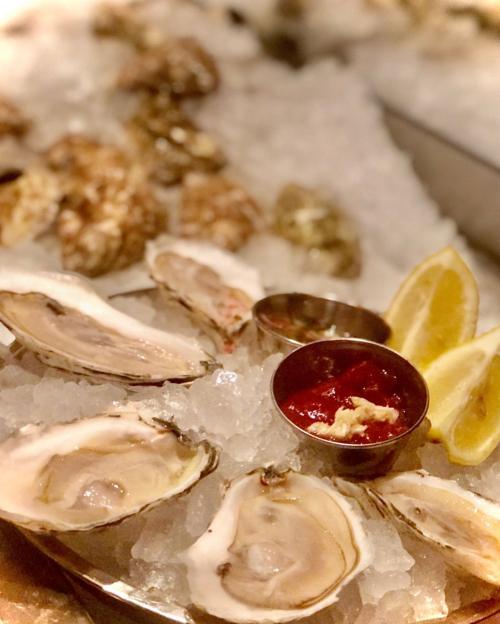 A platter of fresh oysters on ice, accompanied by lemon wedges and a small bowl of cocktail sauce.