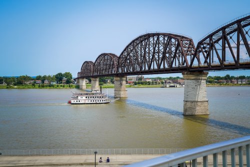 A riverboat sails under a large iron bridge spanning a calm river on a clear day.