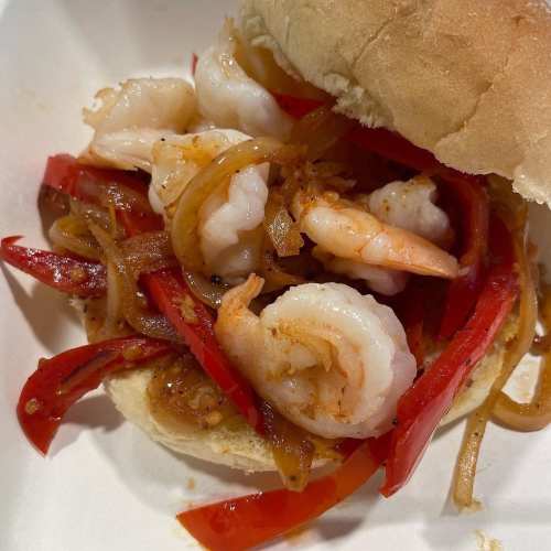 A close-up of a sandwich filled with shrimp, sautéed red peppers, and onions on a soft bun.