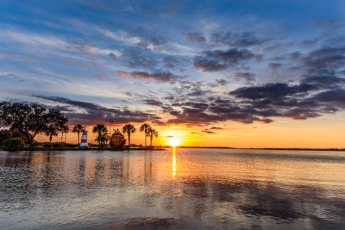 A serene sunset over a calm lake, with palm trees silhouetted against colorful clouds and water reflections.