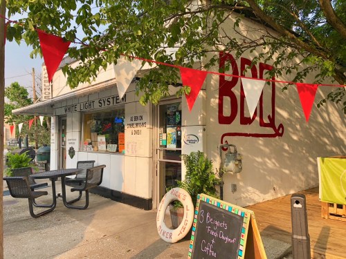 A BBQ restaurant with a red sign, outdoor seating, and festive bunting in a sunny setting.