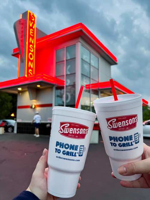 Two hands hold large Swensons cups in front of a brightly lit Swensons drive-in restaurant at dusk.