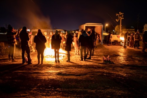 A group of people gathered around a fire at night, with vehicles and lights in the background.