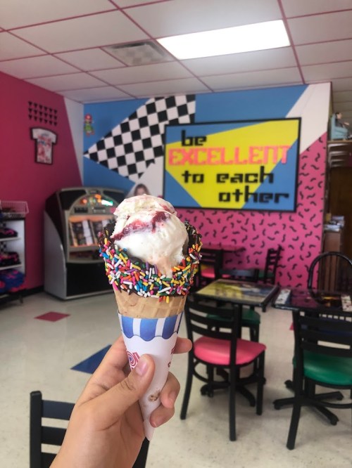 A hand holds an ice cream cone with sprinkles in a colorful ice cream shop featuring a motivational mural.