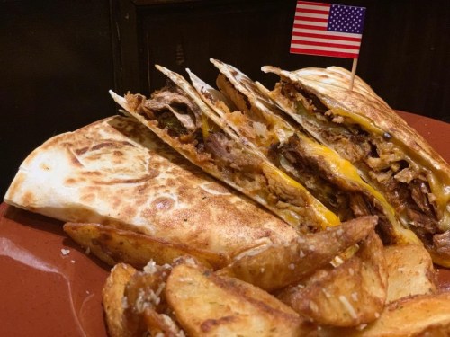 A close-up of a cheesy quesadilla filled with meat, served with potato wedges and a small American flag.