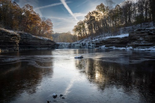 A serene winter landscape with a frozen lake, rocky cliffs, and trees reflecting the soft light of a setting sun.