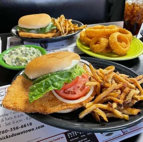 A plate featuring a fried chicken sandwich with lettuce and tomato, fries, onion rings, and a side of green peas.