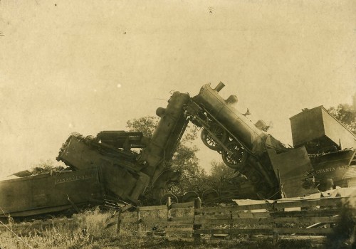 Two derailed vintage steam locomotives leaning against each other in a rural setting, surrounded by overgrown grass.