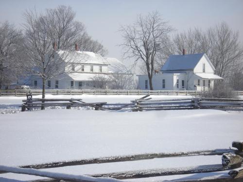 Snow-covered landscape featuring two white houses and bare trees, with a wooden fence in the foreground.