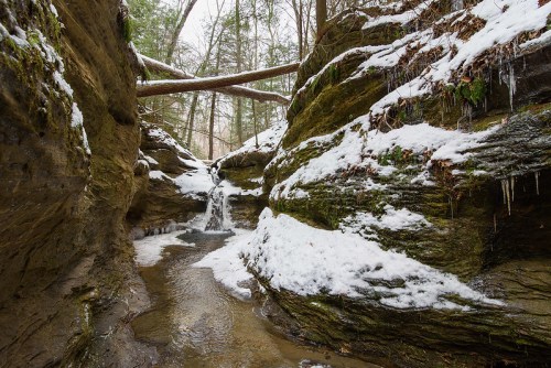 A snowy canyon with a small waterfall, surrounded by steep rock walls and trees in a winter landscape.