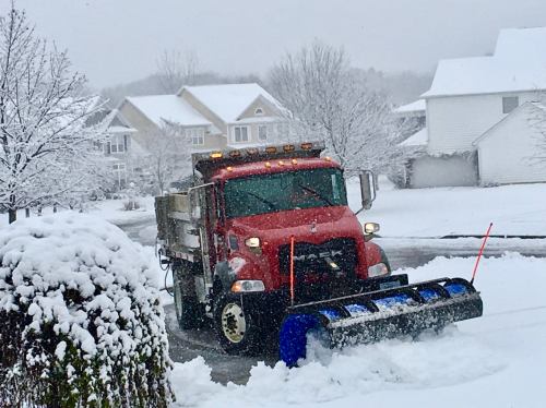 A red snow plow clears a snowy residential street, surrounded by snow-covered houses and trees.
