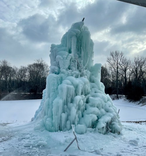 A tall, frozen ice structure with cascading icicles, set against a cloudy sky and bare trees in the background.