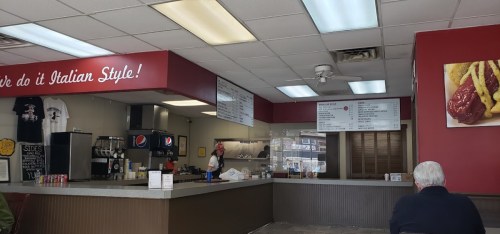 Interior of a casual eatery with a counter, menu boards, and a customer seated. Red and white decor with Italian theme.