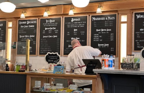 A person prepares orders at a vintage-style ice cream shop with a menu board displaying various treats and drinks.