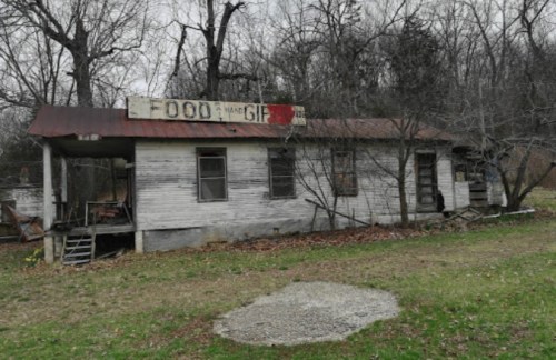 Abandoned building with a weathered sign reading "FOOD" and "HAND GIFT," surrounded by overgrown grass and trees.