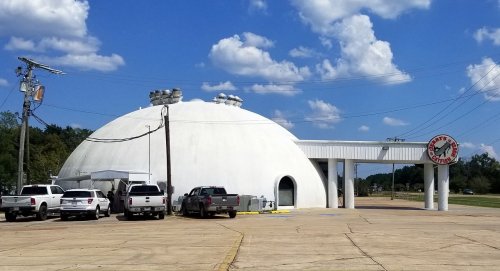 A large white dome-shaped building with a flat roof, surrounded by vehicles, under a blue sky with fluffy clouds.
