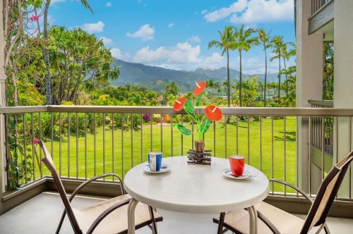 A balcony with a table and chairs, featuring colorful cups and a flower vase, overlooking a lush green landscape and mountains.