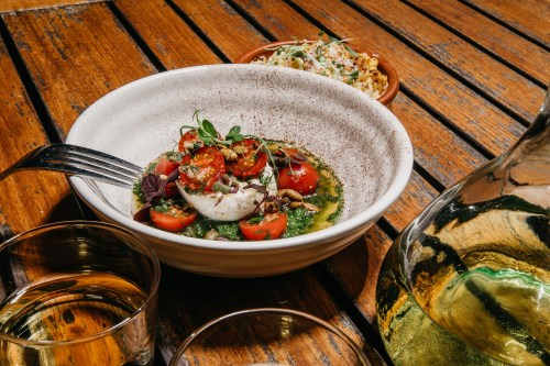 A bowl of burrata with cherry tomatoes and herbs, accompanied by a small dish and a glass of white wine on a wooden table.