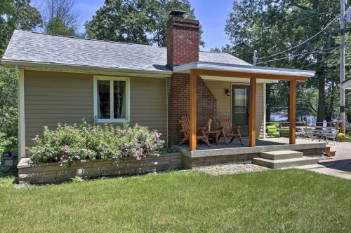A cozy house with a porch, surrounded by greenery and flowers, featuring a chimney and outdoor seating area.