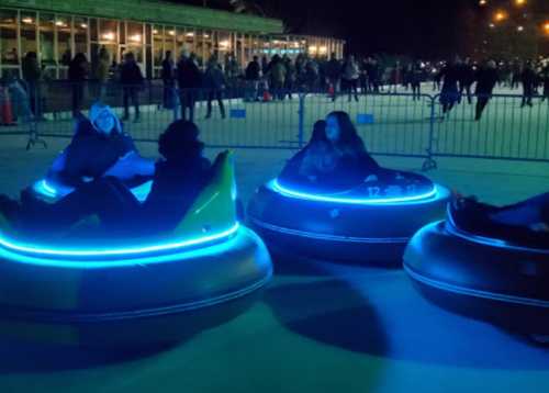 Three people in illuminated bumper cars at an outdoor ice rink, with a crowd and buildings in the background.