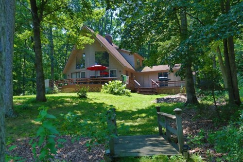 A cozy house surrounded by trees, featuring a deck with an umbrella and a small wooden bridge in the foreground.