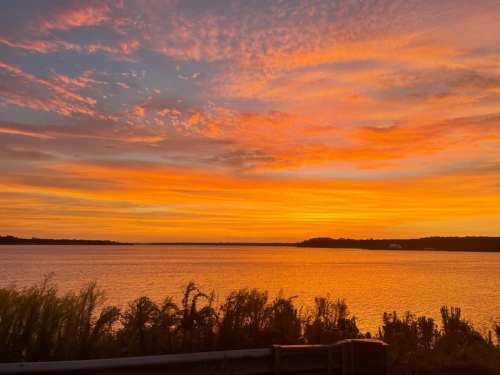 A vibrant sunset over a calm lake, with colorful clouds reflecting on the water and silhouettes of plants in the foreground.