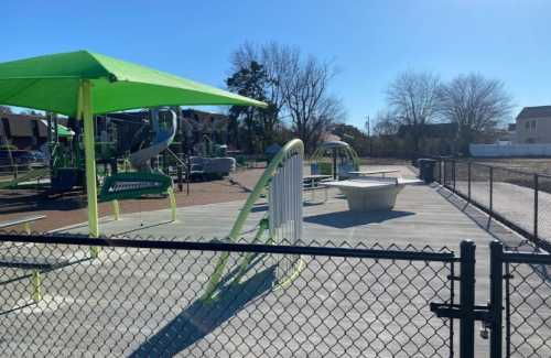 A sunny playground with green equipment, slides, and a fenced area, featuring a picnic table and open space.