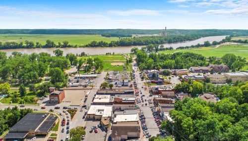 Aerial view of a small town with a river, green fields, and a main street lined with shops and trees.