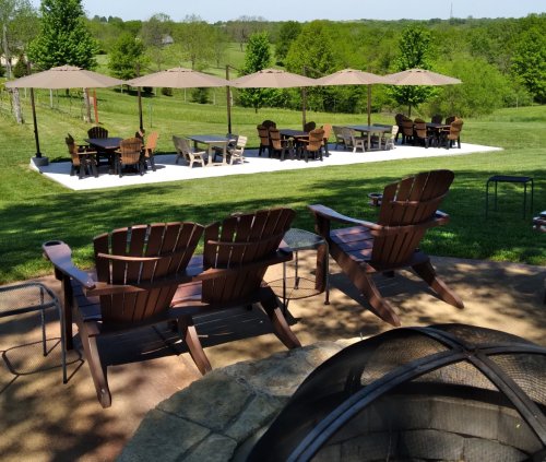 A scenic outdoor area with wooden chairs, tables under umbrellas, and a fire pit, surrounded by green trees and grass.